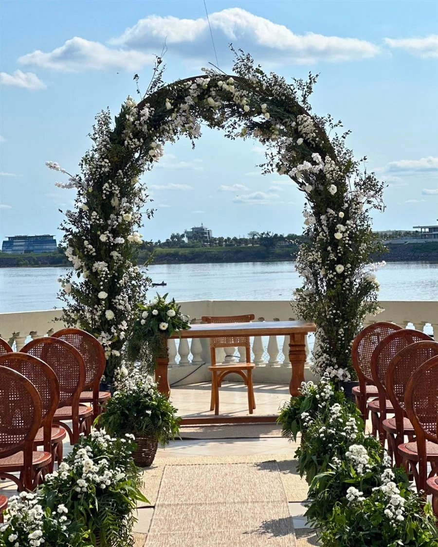 Arco floral blanco frente al río, enmarcando una ceremonia con sillas de mimbre y mesa de madera